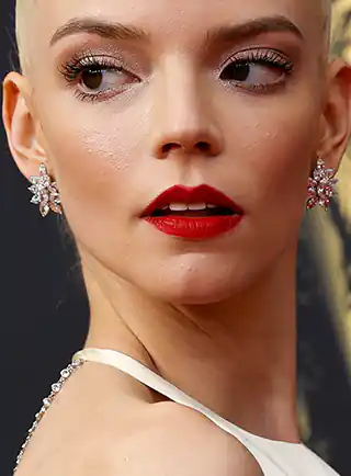 Anya Taylor-Joy is seen against a dark grey and yellow gold background. She wears a pair of large glittery diamond earrings and a deep red matt lipstick. Anya turns towards the camera with her hair tied up in a bun.