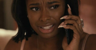 Coco Jones holds a mobile phone to her ear. Her black hair is down and is loose. She wears a dark lipstick with long gel nails and a single large ring in a dark pink bedroom.