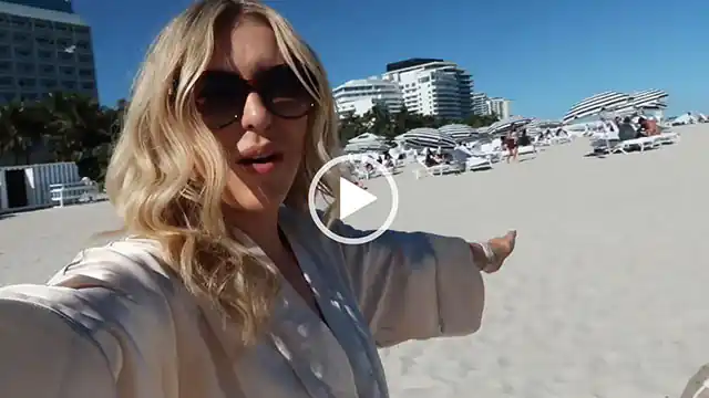 Em Sheldon gestures to the sunlight sandy Miami beach she is walking on. Em wears a gold silk-satin dressing gown with a pair of large black sunglasses. Her blonde hair is loose and curled around her shoulders.