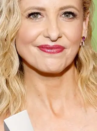 Sarah Michelle Gellar appears in front of a green wall. She has her blonde hair in messy waves. Sarah is seen in a glossy dark pink lipstick and small gemstone earrings. She holds a silver award in frame.