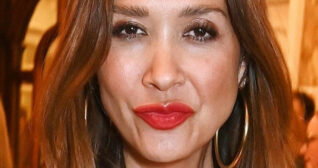 Myleene Klass purses her lips. She wears a bright red lipstick and a pair of large thin gold hoop earrings. Myleene has her long brown hair down as she stands in front of in dark wood and orange wall room.