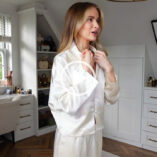 Victoria Magrath adjusts the collar of the white LilySilk pyjama top she is wearing. She stands in her dressing room at home. Victoria has her blonde tone hair loose and down and has on fresh makeup.