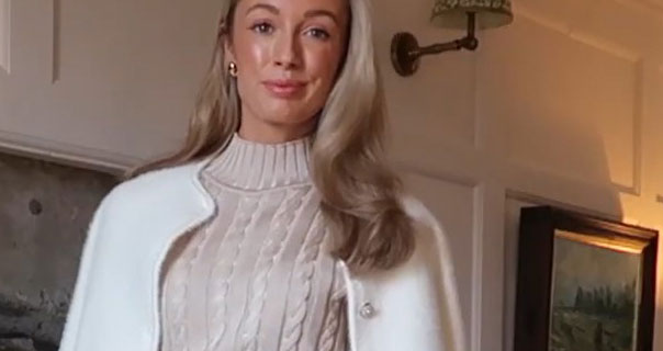 Josie Fear smiles as she models an outfit. She wears a silk thread knitted jumper with a white coat. Josie appears against a white wooden panel wall with stone and a painting behind her.