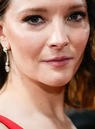 Morfydd Clark is seen with long brown hair that is loose and in waves. She wears a pink slightly sheeny lipstick, black mascara and silver jewel pendant earrings. Morfydd stands in front of a dark backdrop in a red gown.