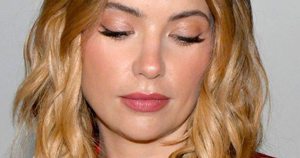 Ashley Benson looks down and to one side with her eyelids almost closed. She wears her blonde hair loose and in waves. She is seen in a red satin blazer against an off-white wall.