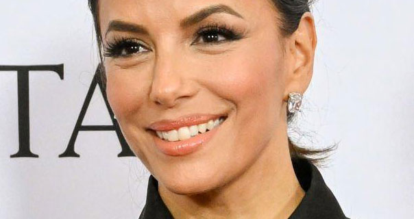 Eva Longoria smiles and looks to the side. She wears a large crystal earring with her hair tied back. She appears in a black shirt in front of a dark white background.