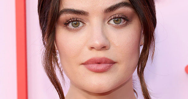 Lucy Hale looks straight ahead. She wears her hair tied up with two strands framing her face. Lucy is seen in pink lipstick against bright background.