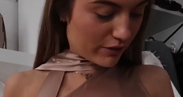 Grace Foley looks downwards with her eyes closed. She wears a copper bronze satin top with a gold necklace in front of cream shelves.