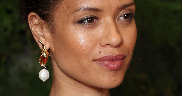 Gugu Mbatha-Raw is seen against a dark black and green background. She wears her hair up with a deep red lipstick and a pair of dangling pearl earrings.