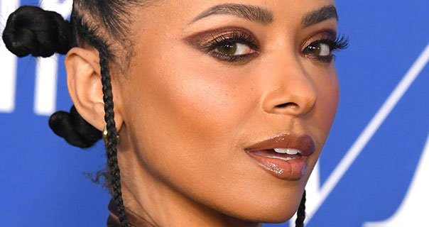 Kat Graham turns to look at the camera. She appears in front of a blue and white background. Her hair is braided and tied in several large knots. She wears brown eye shadow and a glossy lipstick.