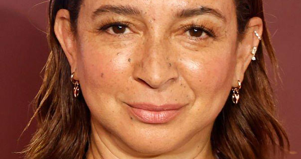 Maya Rudolph is seen in front of a dark red background. Her hair is loose and tucked behind her ears. She wears a deep pink lipstick with gold jewellery.