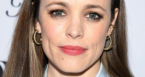 Rachel McAdams is seen in front of a white background that has black text and logos on it. She wears her brunette hair down and back. Rachel appears with red lipstick and a pair of large gold hoop earrings.