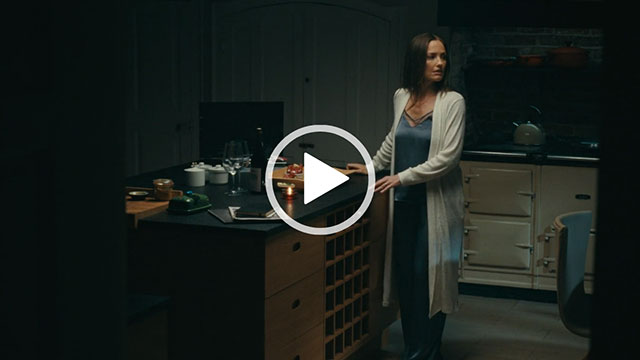 Minka Kelly turns and looks to the side while standing in an open plan kitchen. She wears her hair straight and loose. Minka appears in a long light grey wrap and a pair of dark blue pyjamas.