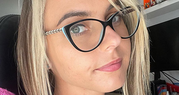 Chloe Toy is seen wearing a pair of large black frame glasses. She wears her short blonde hair down with light makeup and a pink lip.