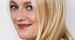 Dakota Fanning is seen turning to look into the camera. She smiles with her blonde hair straight and loose framing her face. She appears in front of an off-white wall with a dark red lipstick and black mascara.