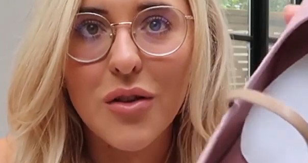 Em Sheldon is seen in the kitchen of her Fulham flat. She wears a pair of large rose gold frame glasses with her hair loose. Em holds up a pink shoe as she sits in front of the black doors that lead into her garden.
