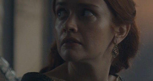 Olivia Cooke looks over her shoulder. She wears her hair braided and tied back as the character Alicent Hightower . She stars in a pair of gold earrings and dark green dress in a room within the Red Keep.