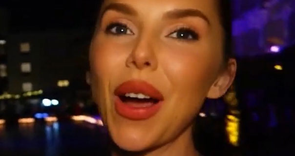 Laura Blair is seen at night-time. She wears a red lipstick and mascara as she records herself in front of a pool lit by orange and purple lamps.