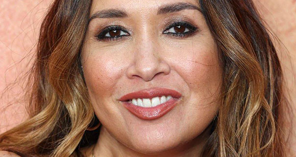 Myleene Klass is seen against a pale orange and pale red background. She has blonde highlights in her brown hair which is loose and slightly curled. Myleene wears a dark red lipstick and she is smiling.