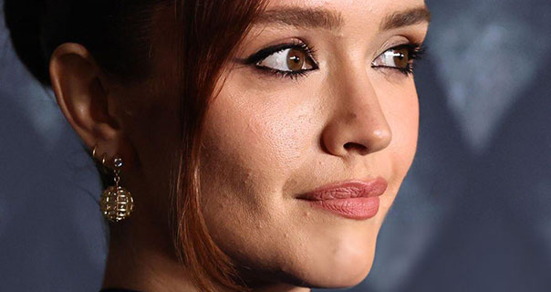 Olivia Cooke looks to the side as she appears in front of a dark blue background. Her hair is tied up and wears gold globe earrings. She has on a dark pink lipstick and a heavy black eyeliner.