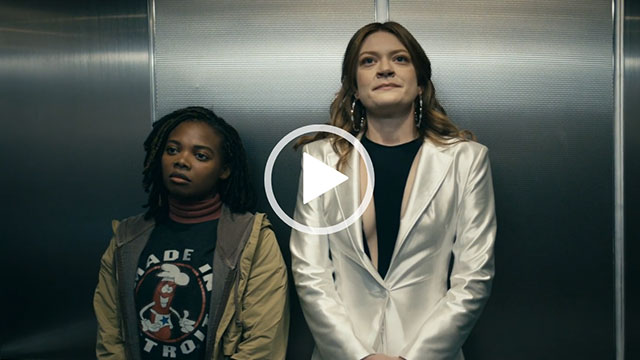 Colby Minifie stands next to Susan Heyward in a metal lift. Colby has her hair loose and wears a pair of large silver earrings. She is seen in a white shiny blazer over a black and beige top.