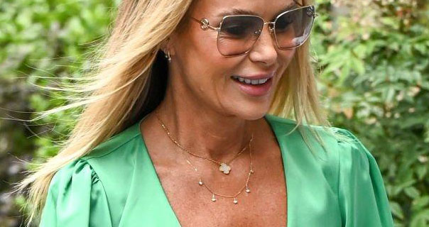 Amanda Holden smiles with her blonde hair down and blowing in the wind. She wears a green plunge top with large metal frame sunglasses and a thin gold necklace.