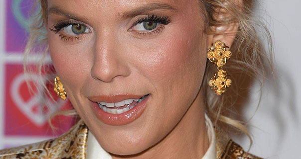 AnnaLynne McCord wears a pair of ornate dangling yellow earrings with her hair tied up. She appears in front of a white, pink and purple wall. AnnaLynne is seen in a shiny blazer with a white nylon shirt.