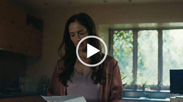 Minka Kelly looks down at a white letter she is holding in both hands. She wears a dark pink satin dressing gown and grey sleepwear. Her long black hair i loose and slightly messy. Minka stands in a cream colour kitchen in front of a large window with greenery just outside in dappled sunshine.