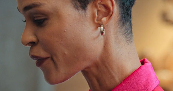 Godeliv Van den Brandt is seen in side profile. She wears a pair of gold circle earrings with her black hair cut short. Godeliv appears in a structured pink shirt a grey and dark yellow room.