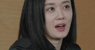 Jang Na-ra is seen standing and leaning forward in front of a light brown wall. She wears a long sleeve shirt with her shoulder length short black hair down and straight.