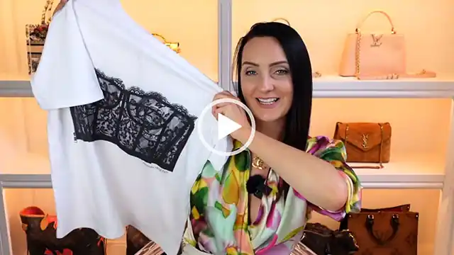 Steph Turton holds up a white t-shirt with a black corset print. She wears a green, pink and yellow wrap-over top. Steph sits in front of her handbag filming display of white shelves with pale orange lighting.