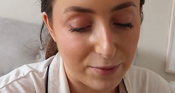 Hazel Maria Wood wears a white pyjama top as she sites in bed. She is seen in front of her grey headboard and cream walls. Hazel has her light brown hair tied back and wears makeup and a dark pink lipstick.