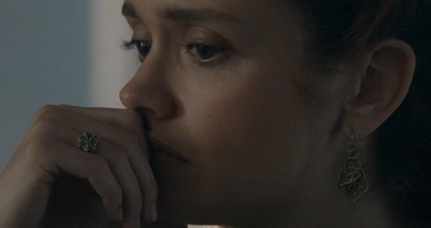 Olivia Cooke touches her face with her hand as she thinks. She wears a gold lattice ring with gold kite shape earrings. Olivia is seen from the side as she sits in front of a blue colour background.