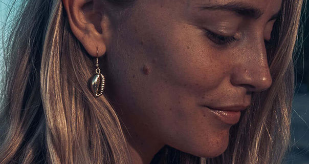 A side profile of Janni Delér. Janni has her eyes closed as she appears on a beach as the sun begins to set. She wears a pair of dangling gold shell earrings with her blonde hair loose.