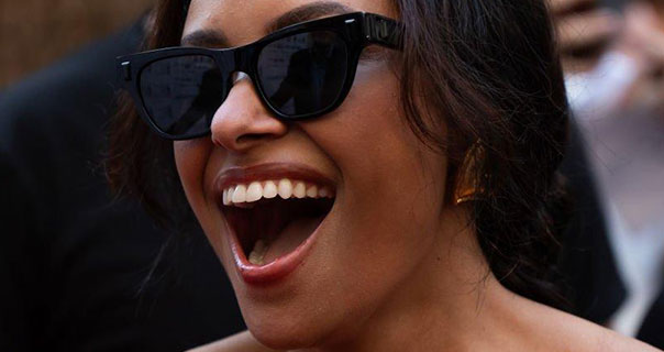 Kat Graham is seen wearing a pair of large black sunglasses with her mouth open wide. She wears a dark red glossy lipstick as she appears in front of fans.