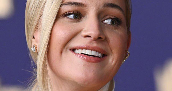 Kelsea Ballerini smiles and looks to the side. She has her blonde hair straight and tied back. Kelsea wears a pink gloss lipstick and jewel earrings as she stands by a purple and gold wall.