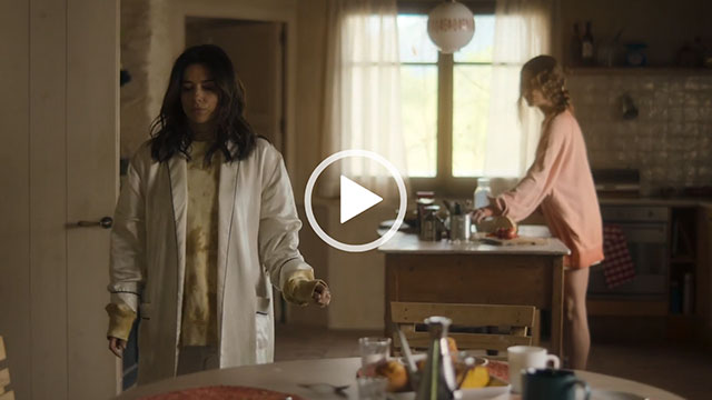 Eva Longoria walks through a white wood kitchen. She makes her way to a large circular table. Eva wears a long white satin dressing gown over a faded yellow jumper.