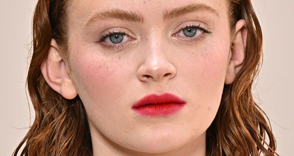 Sadie Sink stands in front of a beige background with a straight expression. She wears her hair down and slicked back. Sadie wears a dark red matte lipstick.