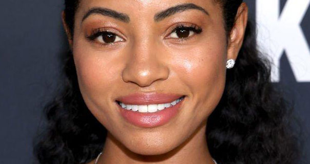 Camille Hyde smiles as she stands in front of a dark blue and white background. She wears a rich gloss lipstick and jewel stud earrings with her black hair loose, swept back and curled.