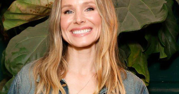 Kristen Bell is seen in front of a large dark green leaf plant. She smiles widely with her blonde hair loose around her shoulders. Kristen wears a denim jacket and a thin dangling necklace.