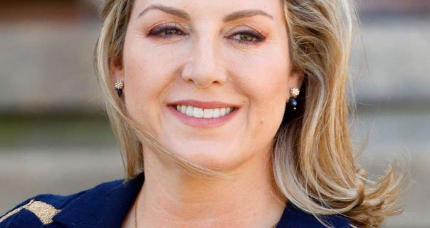 Penny Mordaunt smiles. Her blonde hair is loose and blowing in the wind. She wears a pair of small earrings and a dark pink lipstick with a navy and dark gold cardigan.