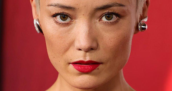 Pom Klementieff looks straight ahead. She is seen in front of a red wall. Pom wears a matt red lipstick, mascara and a pair of large silver earrings with her hair blonde and cut short.