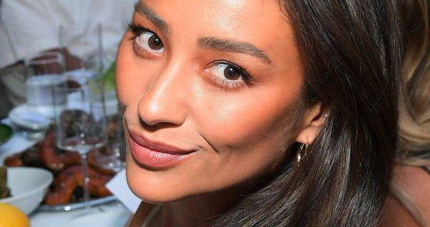 Shay Mitchell turns and tilts her head to one side as she sits at a white table full of food and drinks. Her brown hair is straight and down. She wears a dark pink lipstick and dangling earrings.