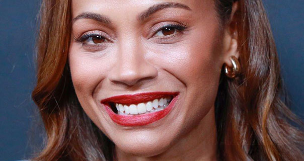 Zoe Saldana smiles widely. She wears a dark red lipstick and small chunky gold hoop earrings. Zoe appears in front of a dark blue wall with her brown hair loose and slightly curled.