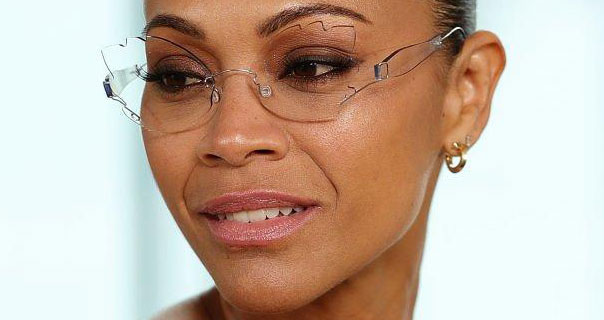 Zoe Saldana wears a pair of frameless see through glasses. She has her black hair up. Zoe appears in small gold hoop earrings and a dark pink gloss lipstick in front of a light blue background.