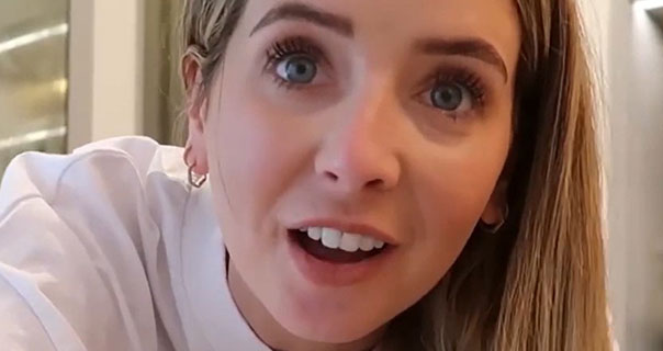 Zoe Sugg peers into the camera. She appears in her large white and cream dressing room at home. Zoe wears her long brown hair loose and down. She is seen in small gold earrings and a white t-shirt in front of full length wardrobe mirrors.