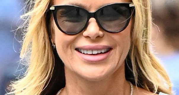 Amanda Holden smiles with her blonde hair loose and slightly curled. She wears a pair of large sunglasses with a thick matt light pink lipstick and a silver necklace.