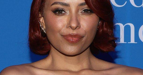 Kat Graham looks into the camera as she stands in front of a blue wall with large white text on it. She wears her hair cut short and tinted red. Kat is seen in a glossy lipstick and large earrings.