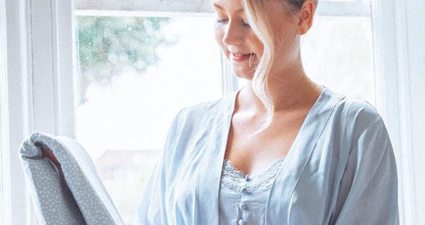 Meg Says is seen in front of a large white frame window during the day. She has her blonde hair tied up in a bun with loose strands framing her face. Meg wears a baby blue dressing gown and delicate blue pyjamas.