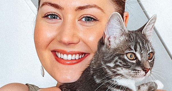 Meg Says holds and cuddles her cat Millie. She has her hair up in a bun and wears a glossy lipstick with pendant earrings. Meg is seen in front of the white staircase of her cottage home.